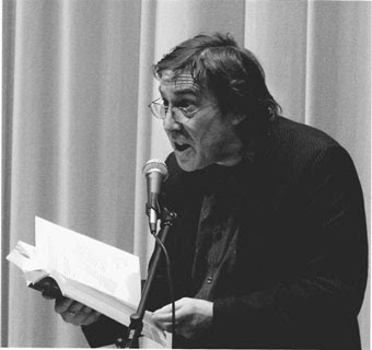 Black and white photograph of Alan Halsey reading from a book into a microphone.