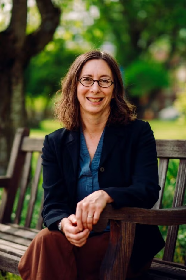 Color photograph of Adela Pinch sitting on a park bench in front of a tree.