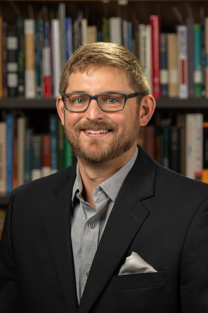 Color photograph of Chet Lisiecki wearing a suit in front of a bookcase. 
