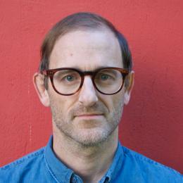 Headshot of Alastair Hunt wearing glasses in front of a red wall. 
