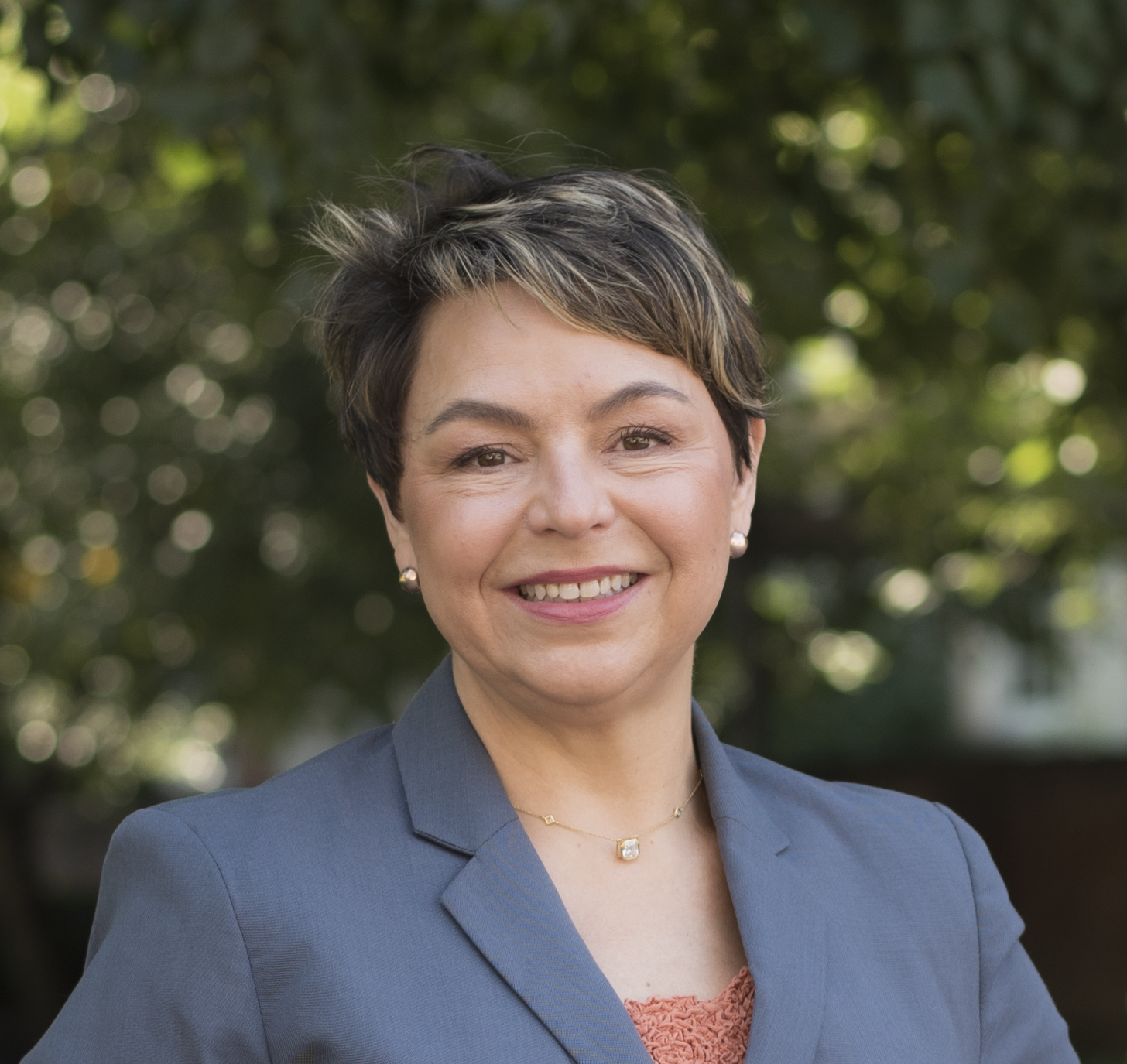 Color headshot of Andrea Rehn in a suit jacket in front of greenery.